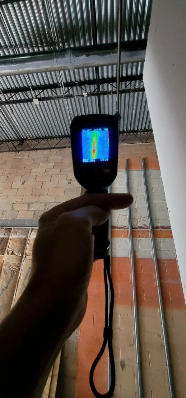 Person holding a thermal camera pointing at a ceiling with metal roofing and exposed pipes.