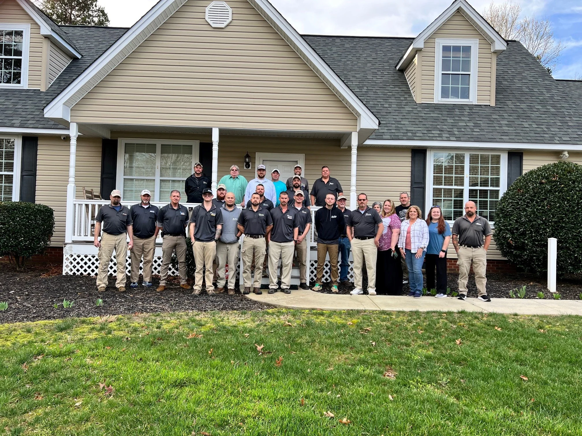 A large group of people standing on the porch and yard in front of a beige house with white trim, black shutters, and a gabled roof, some wearing matching black shirts, others in casual clothing.