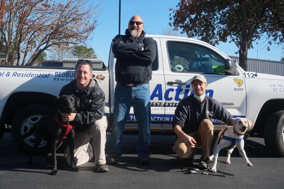 Three men with two dogs are posing in front of a white service truck with blue lettering, outdoors on a sunny day with trees in the background.