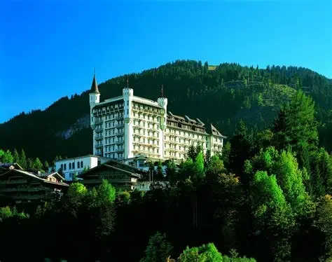A large hotel or castle-like building on a hill surrounded by trees with a forested mountain in the background under a clear blue sky.