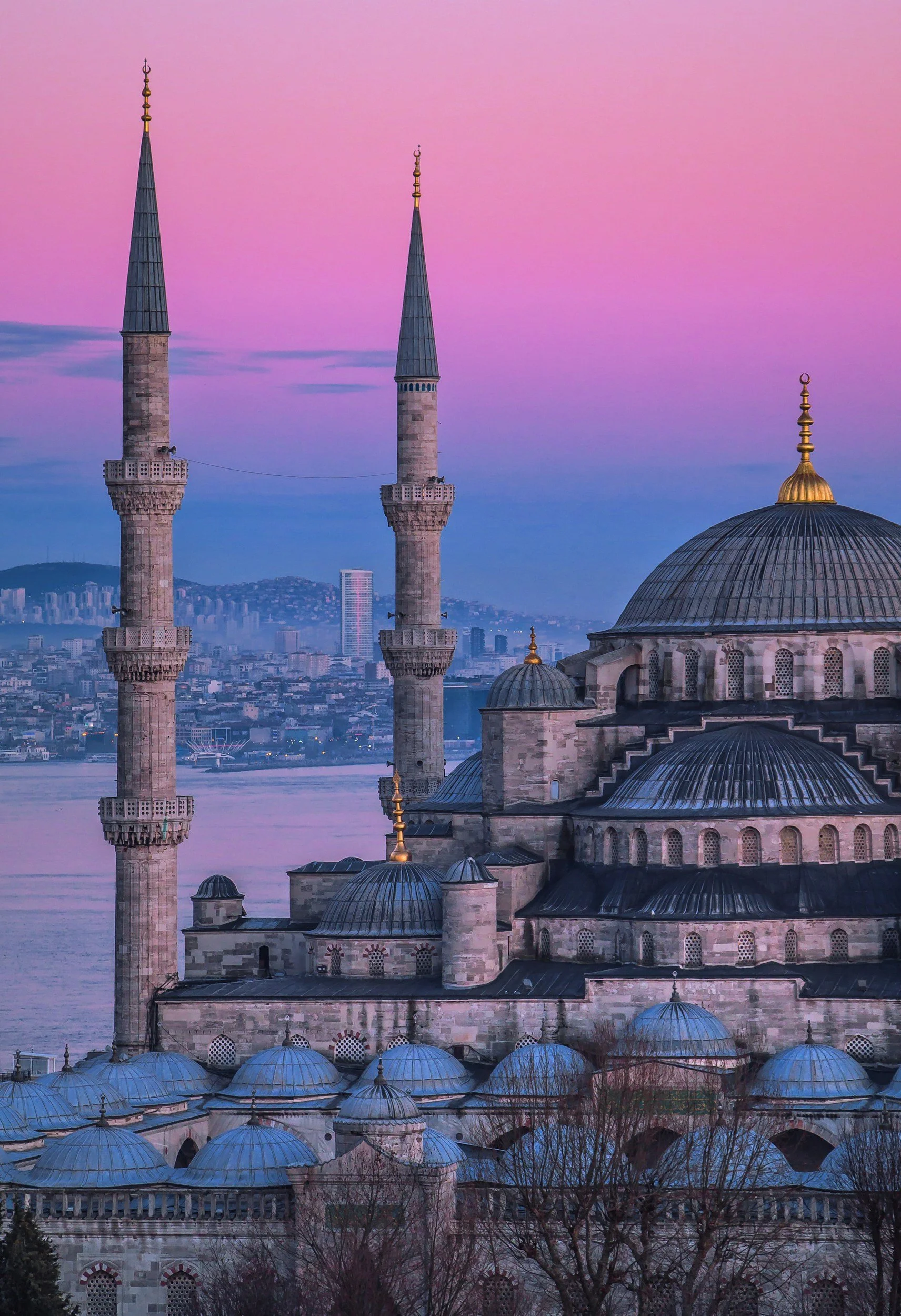 A mosque with multiple domes and two tall minarets, set against a colorful sunset sky over a city and water body in the background.