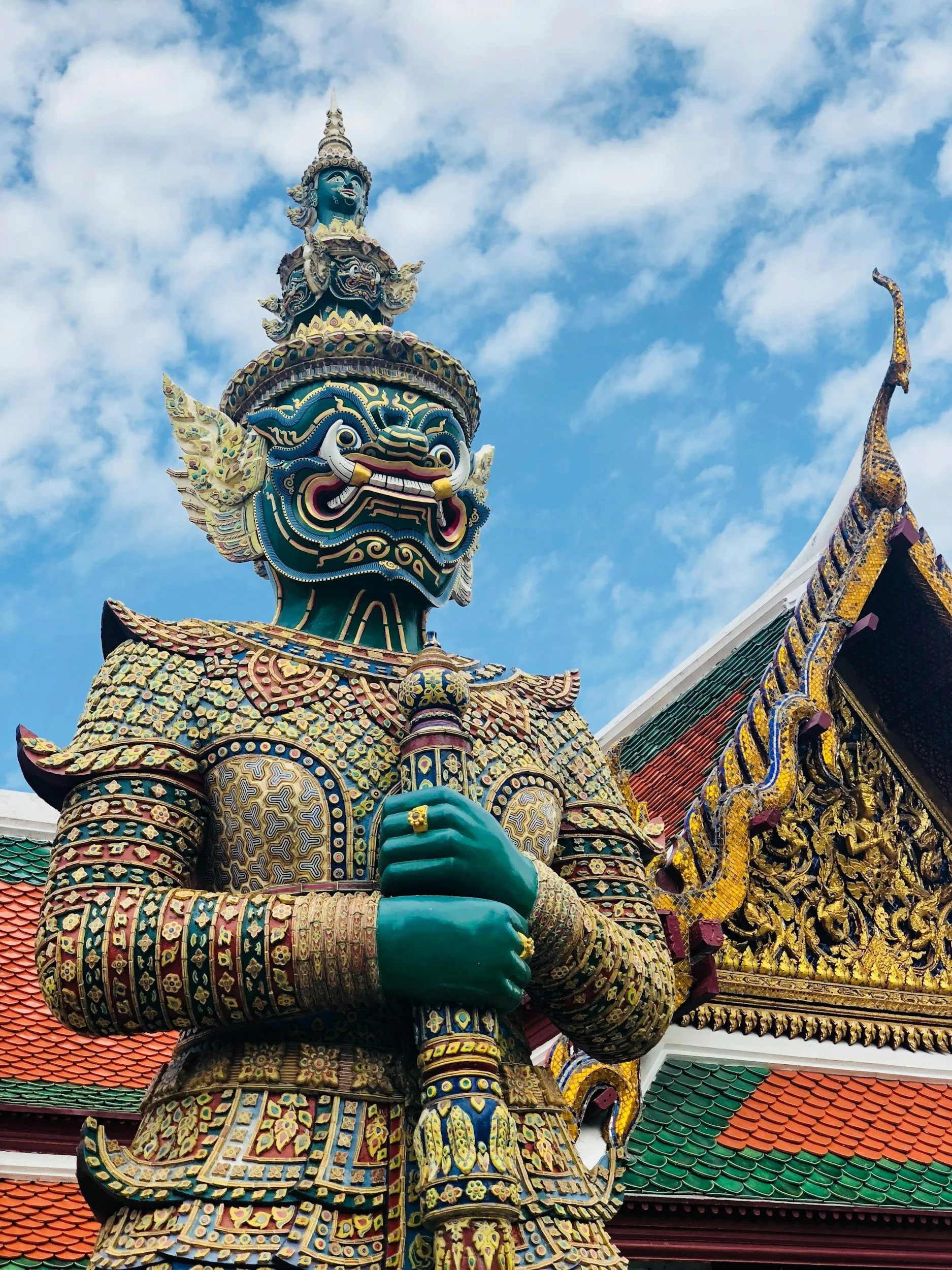 A large colorful statue of a mythological figure standing in front of a temple with ornate architecture and a blue sky background.