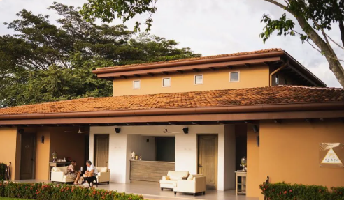 Two people sitting outside a modern house with a large patio, surrounded by greenery and trees.