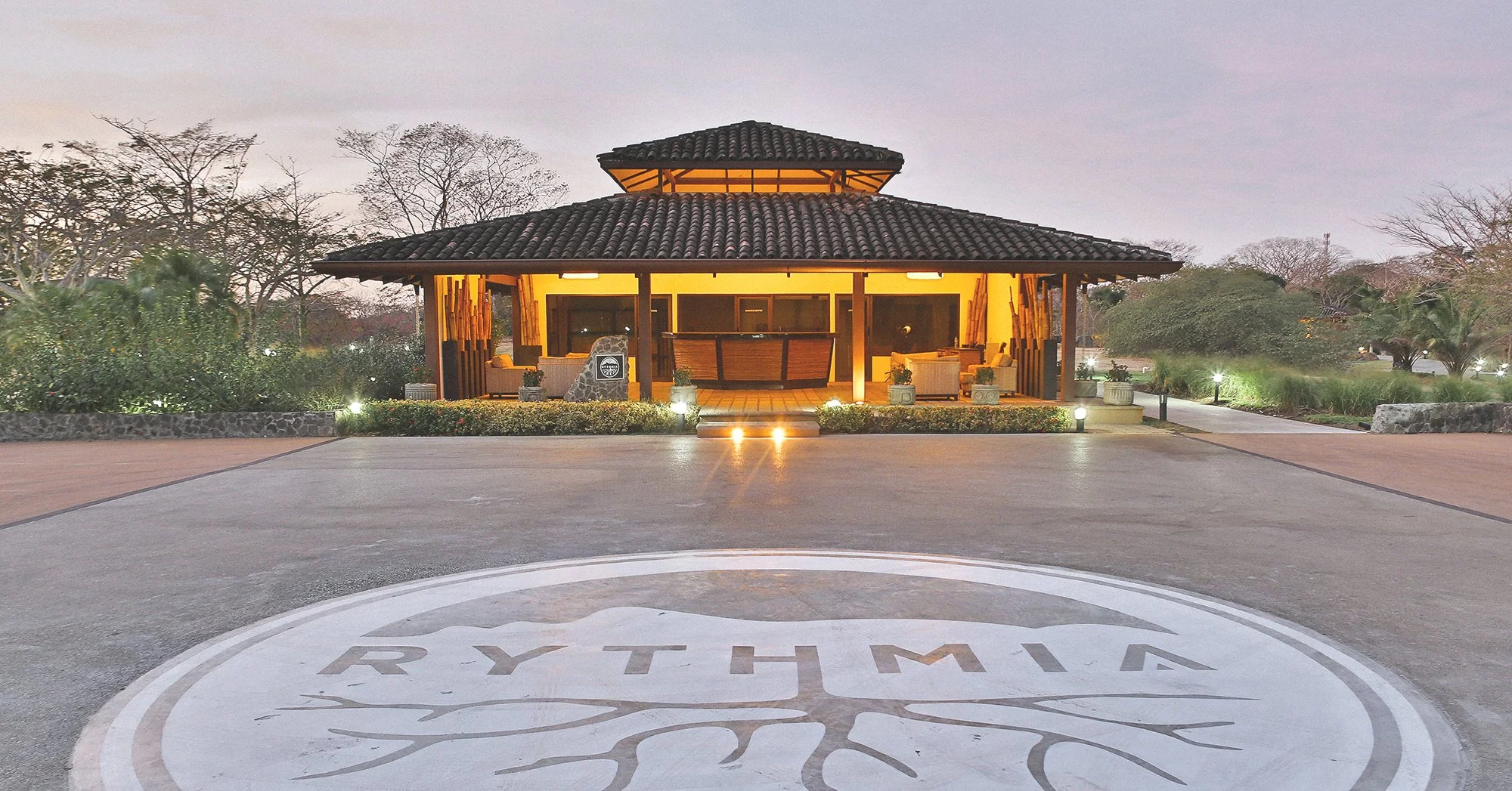 A luxurious outdoor pavilion with warm lighting, surrounded by trees and greenery, with the word 'RYTHMIA' and a tree logo painted on the ground in front.