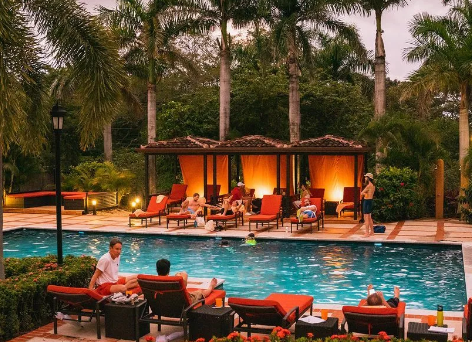 People relaxing on lounge chairs around a swimming pool at sunset with palm trees and cabanas in the background.