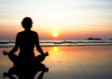 A person practicing yoga or meditating on the beach at sunset with the ocean in the background.