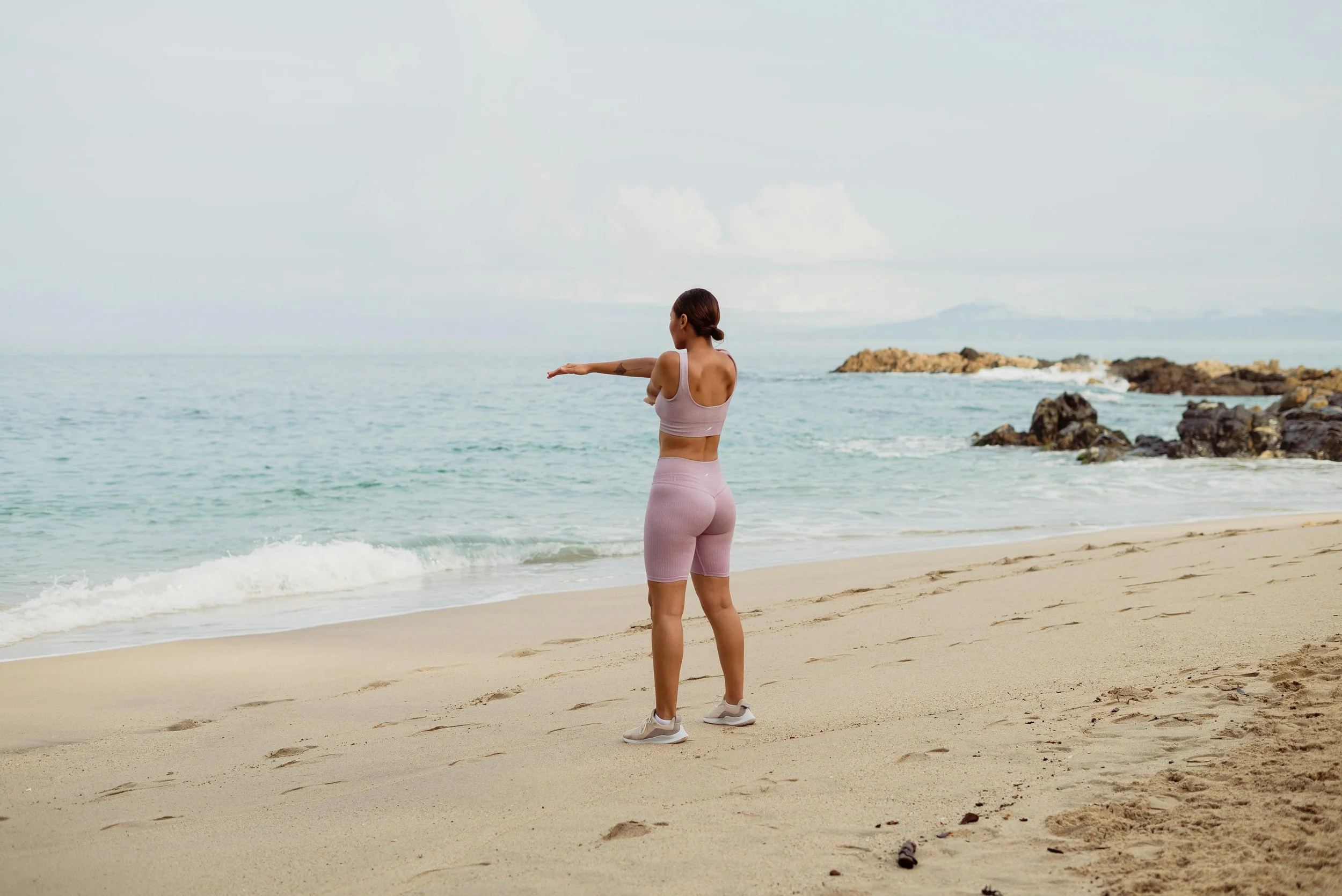 A woman in athletic wear stretching on a sandy beach near the ocean.