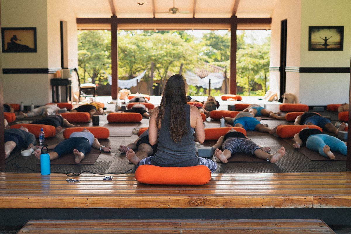A yoga class in a spacious room with large windows, participants lying on mats with orange bolsters, and an instructor sitting on a cushion facing away.