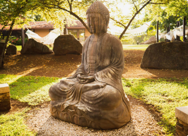 A stone statue of Buddha sitting in a meditative pose outdoors, surrounded by rocks and trees.