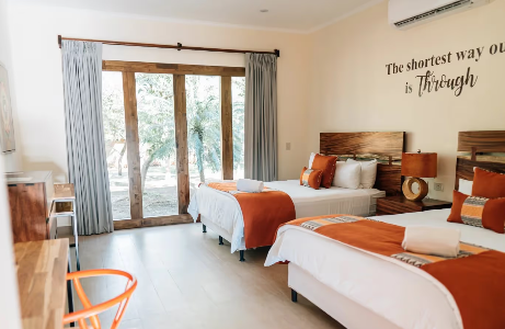 A bright hotel room with two beds, each with orange and white bedding and pillows, wooden headboards, a large window with sliding doors and blue curtains, a small desk, and a wall decal that reads 'The shortest way out is through'.