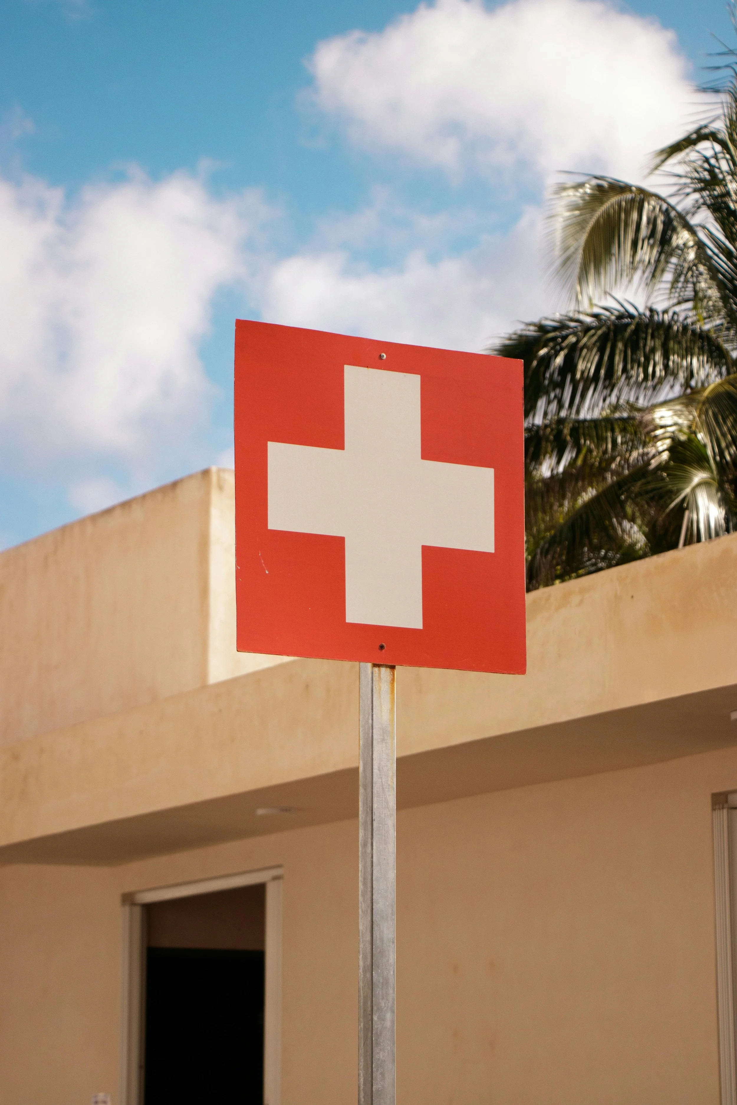 A sign with the red and white Swiss cross symbol outside a building with a beige facade under a blue sky with some clouds and palm trees in the background.