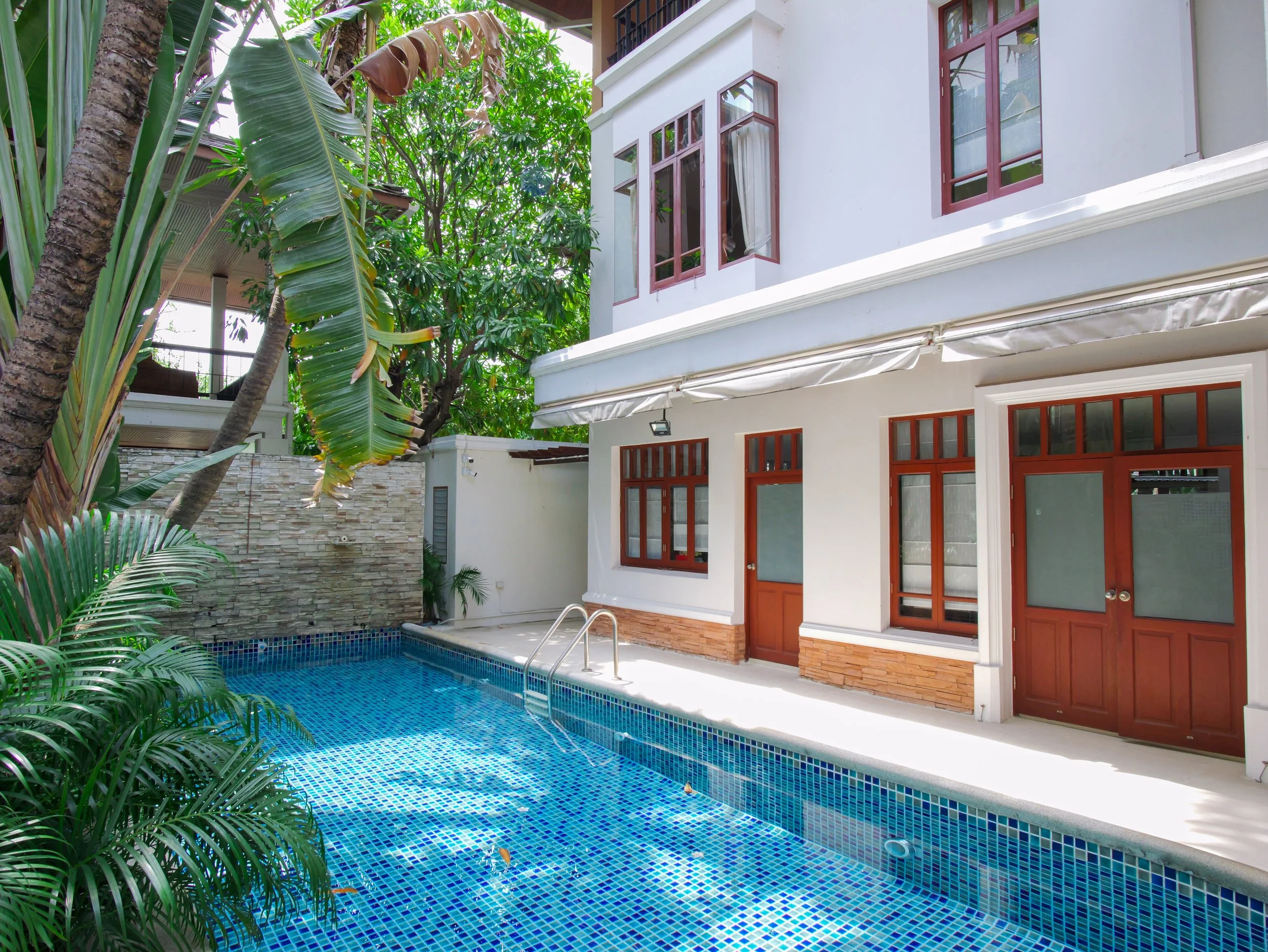 Exterior view of a house with a small swimming pool surrounded by greenery, including palm trees and other tropical plants.