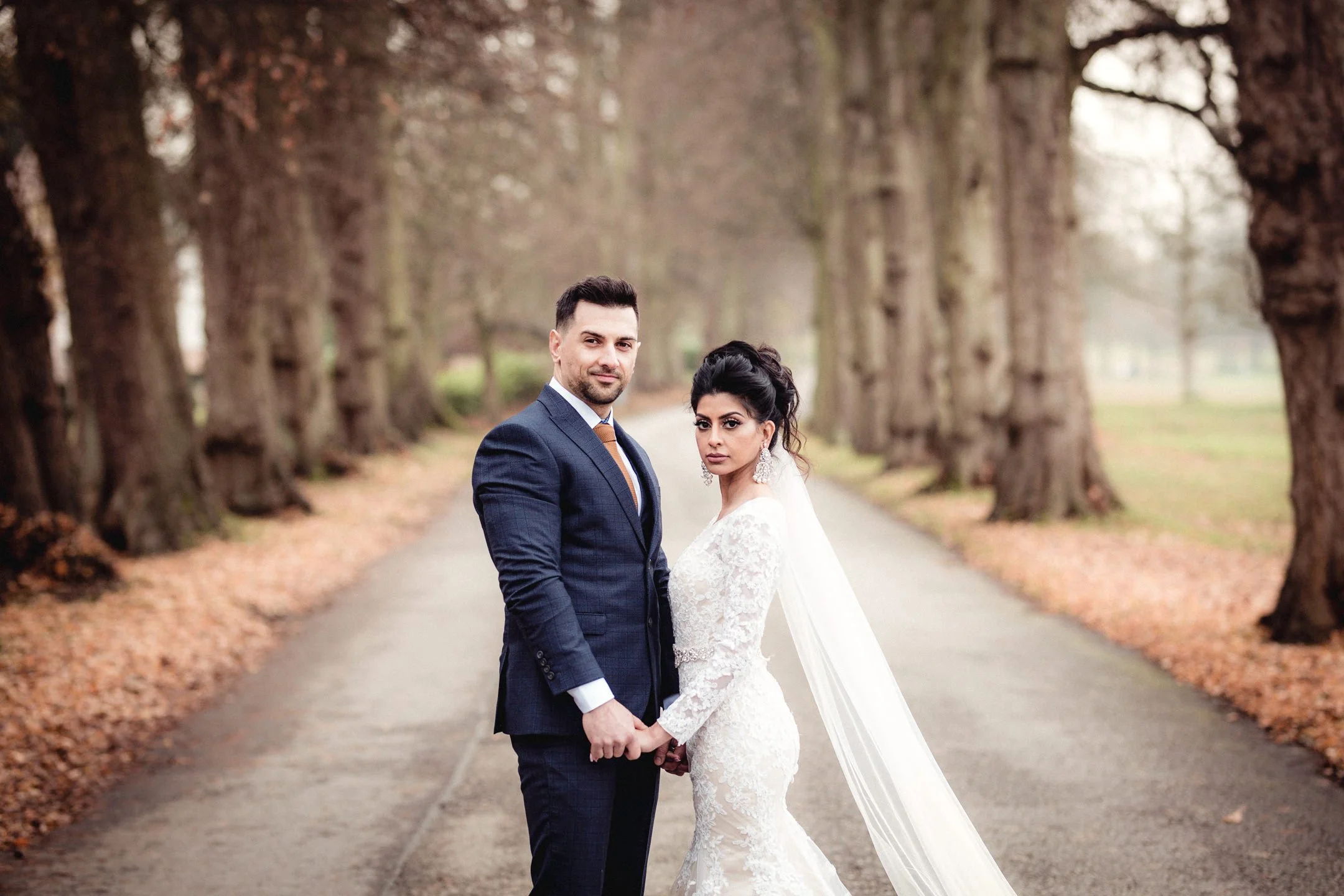 A newlywed couple holding hands stands on a tree-lined path, the groom in a dark suit and the bride in a lace wedding dress with a veil, in an outdoor setting during fall.