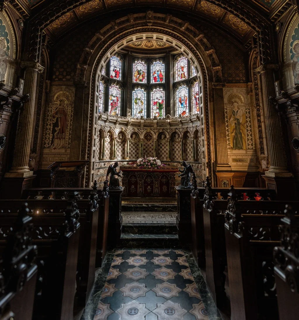 Inside a church or chapel with ornate stained glass windows depicting religious figures, intricate woodwork, and statues, with an altar at the end adorned with flowers.