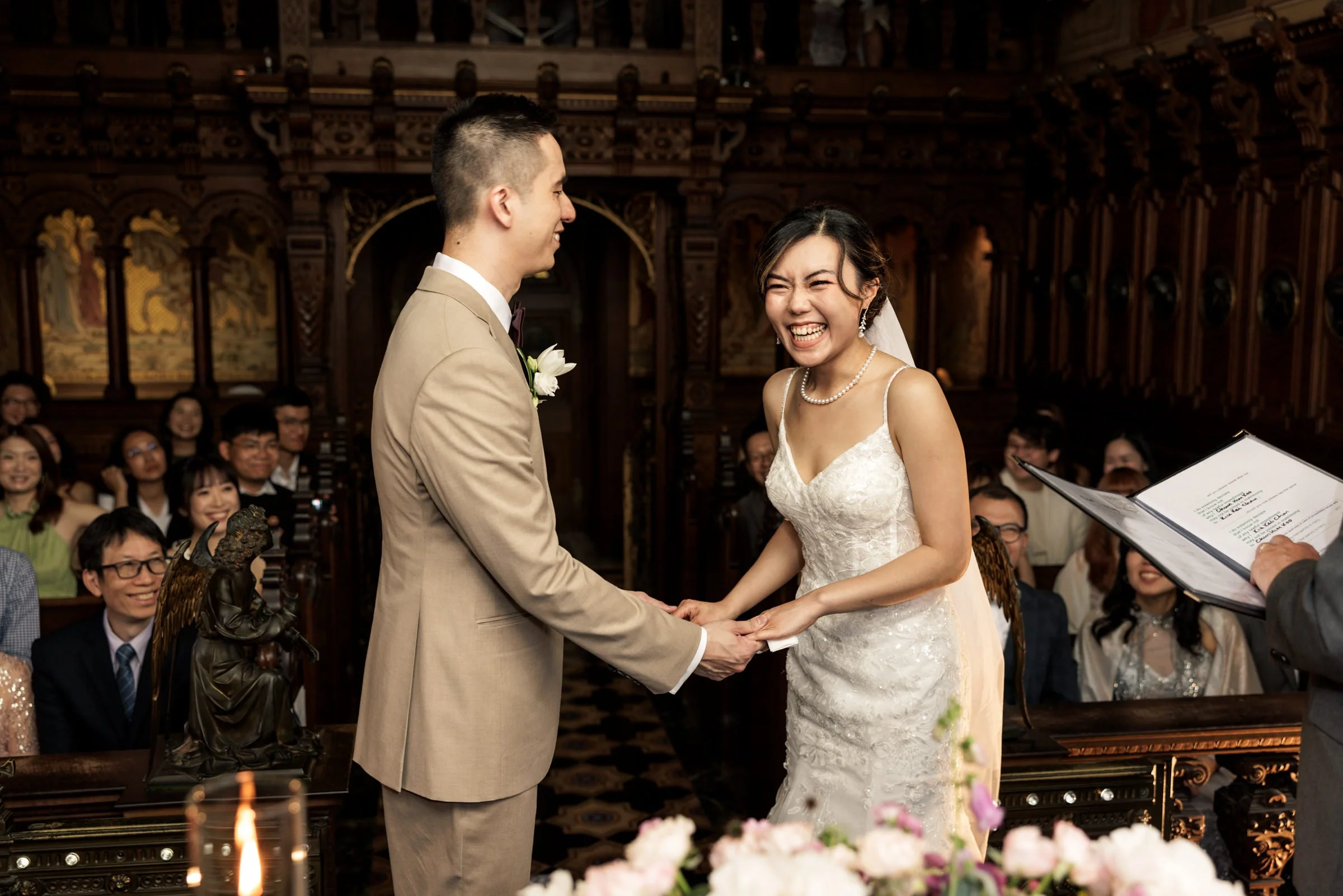 A bride and groom exchange vows during a wedding ceremony inside a church, with wedding guests watching and smiling.
