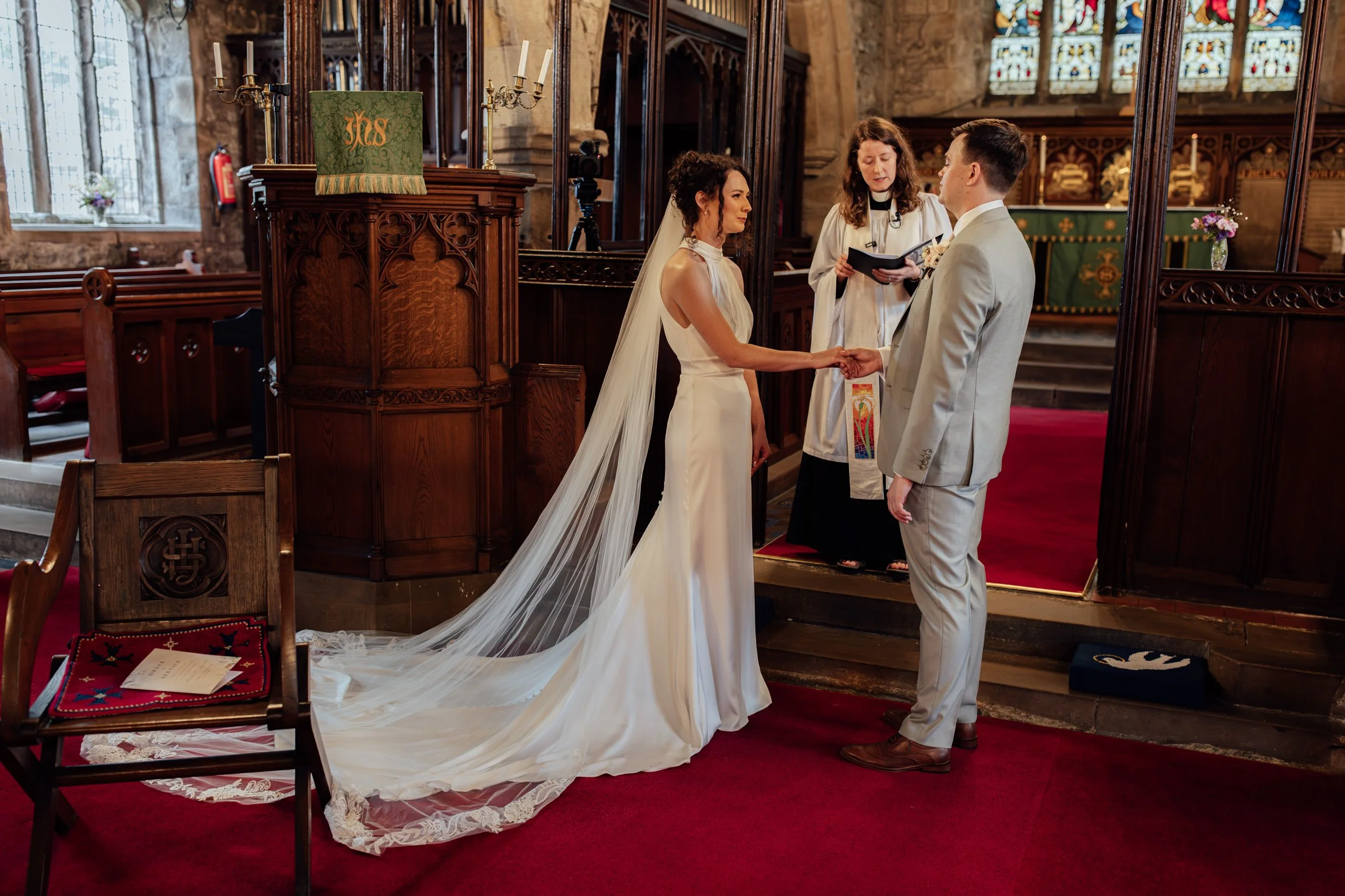 A wedding ceremony inside a church with a bride and groom holding hands before an officiant. The bride is in a white gown with a long veil, and the groom is in a light gray suit. The church has wooden pews, stained glass windows, and red carpet.