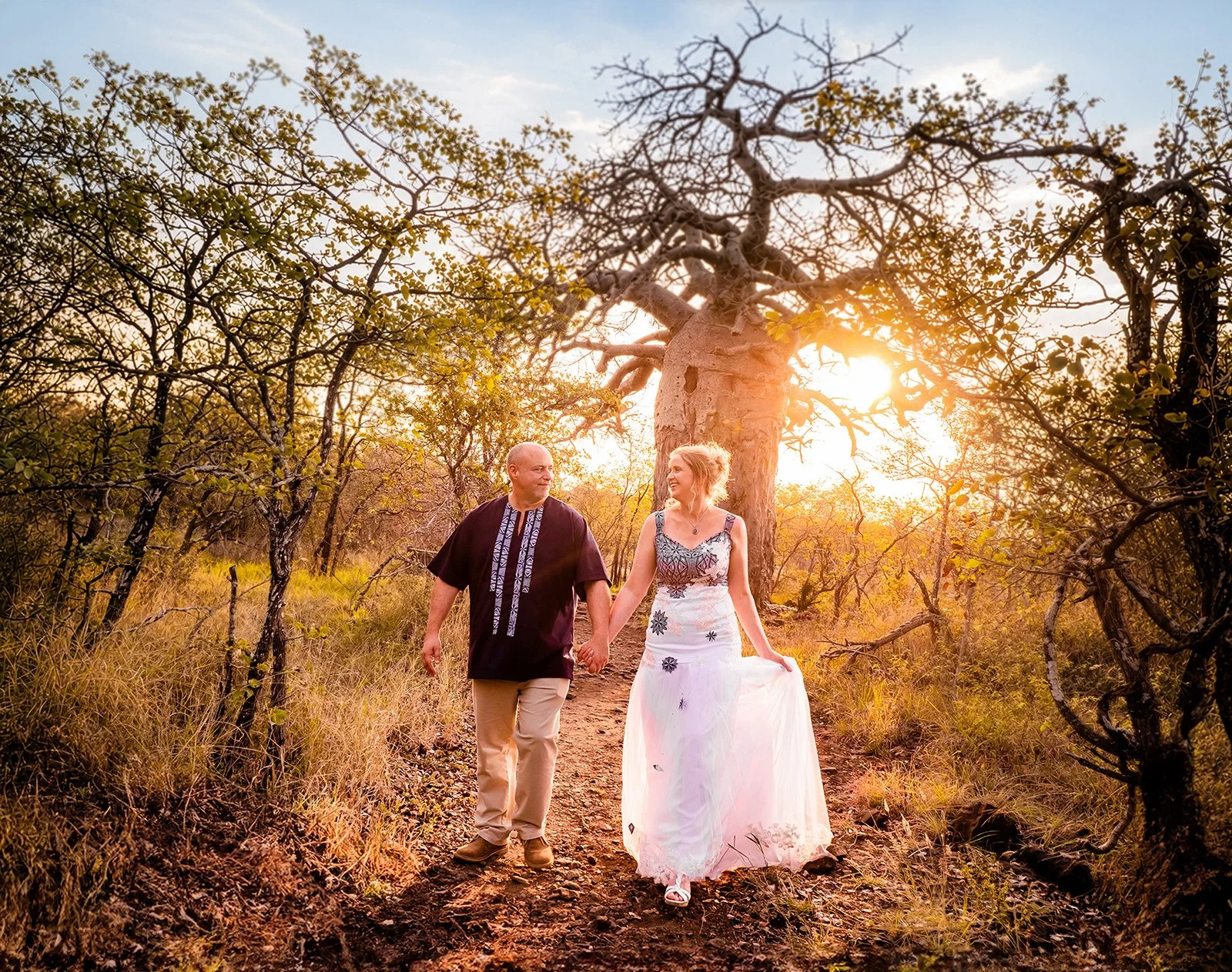 A couple holding hands while walking on a dirt trail through a wooded area at sunset. The woman is wearing a white strapless dress with floral embroidery, and the man is wearing a dark traditional shirt and beige pants. The scene is illuminated by warm golden sunlight filtering through the trees.