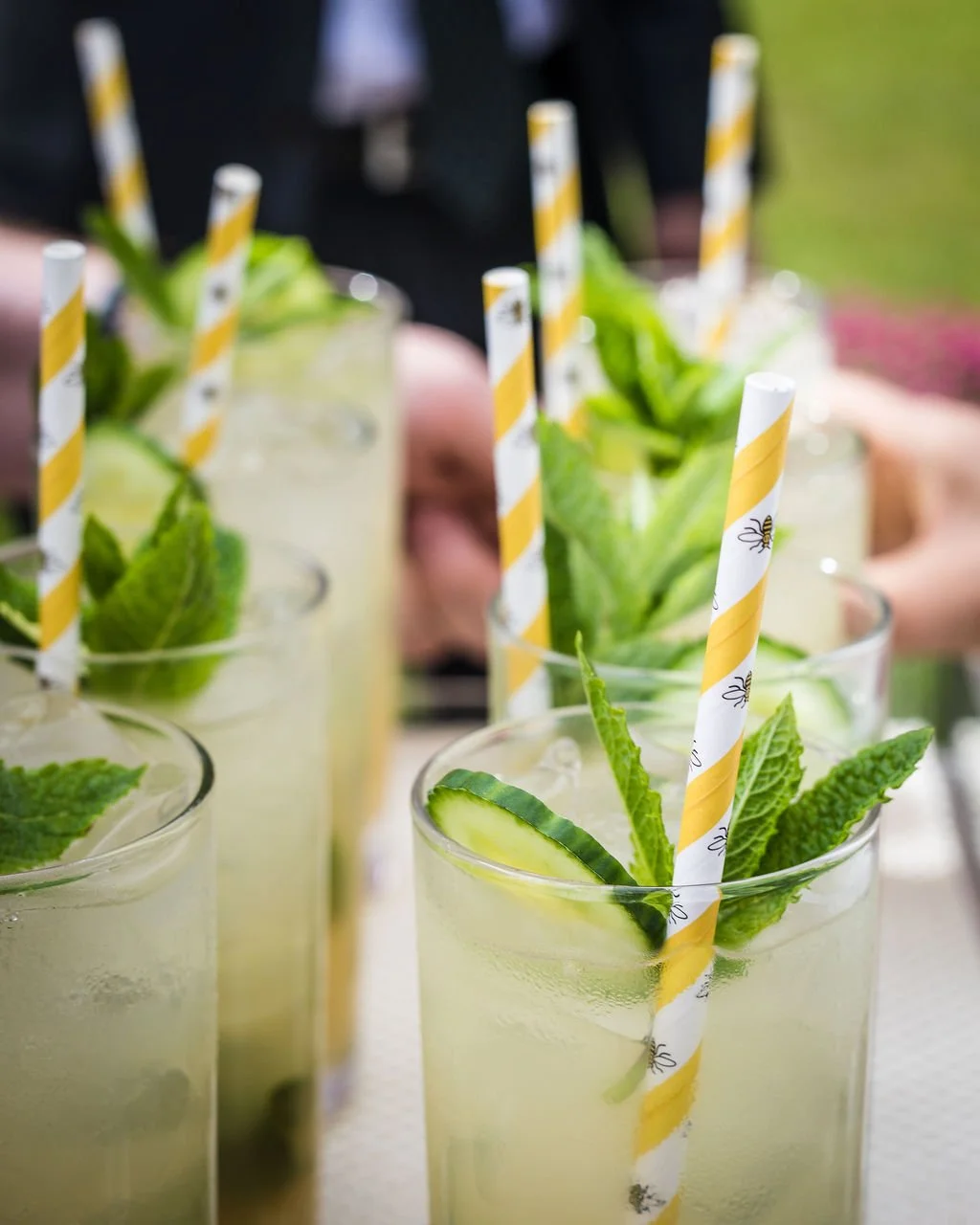 Several glasses of cucumber, mint, and lemon drinks with striped paper straws on a table, outdoors.