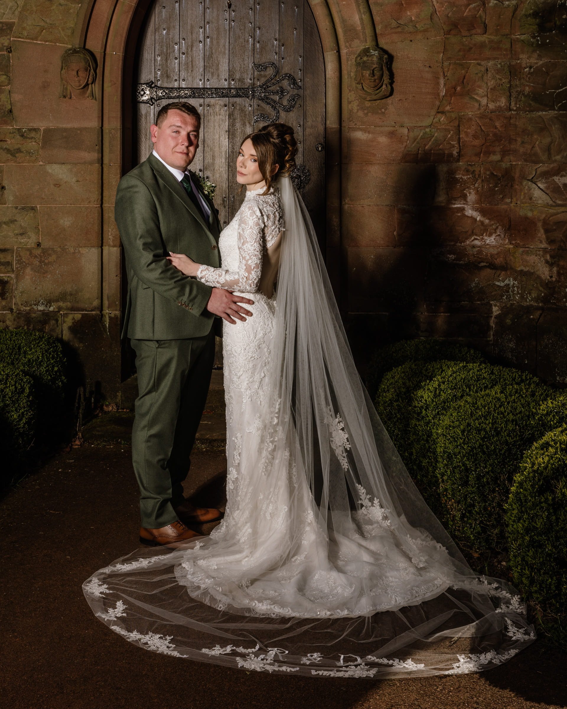 A newlywed couple standing in front of a wooden door with iron hinges, the groom in a green suit and tie, the bride in a lace wedding gown with a long veil, surrounded by a stone wall and well-trimmed bushes.