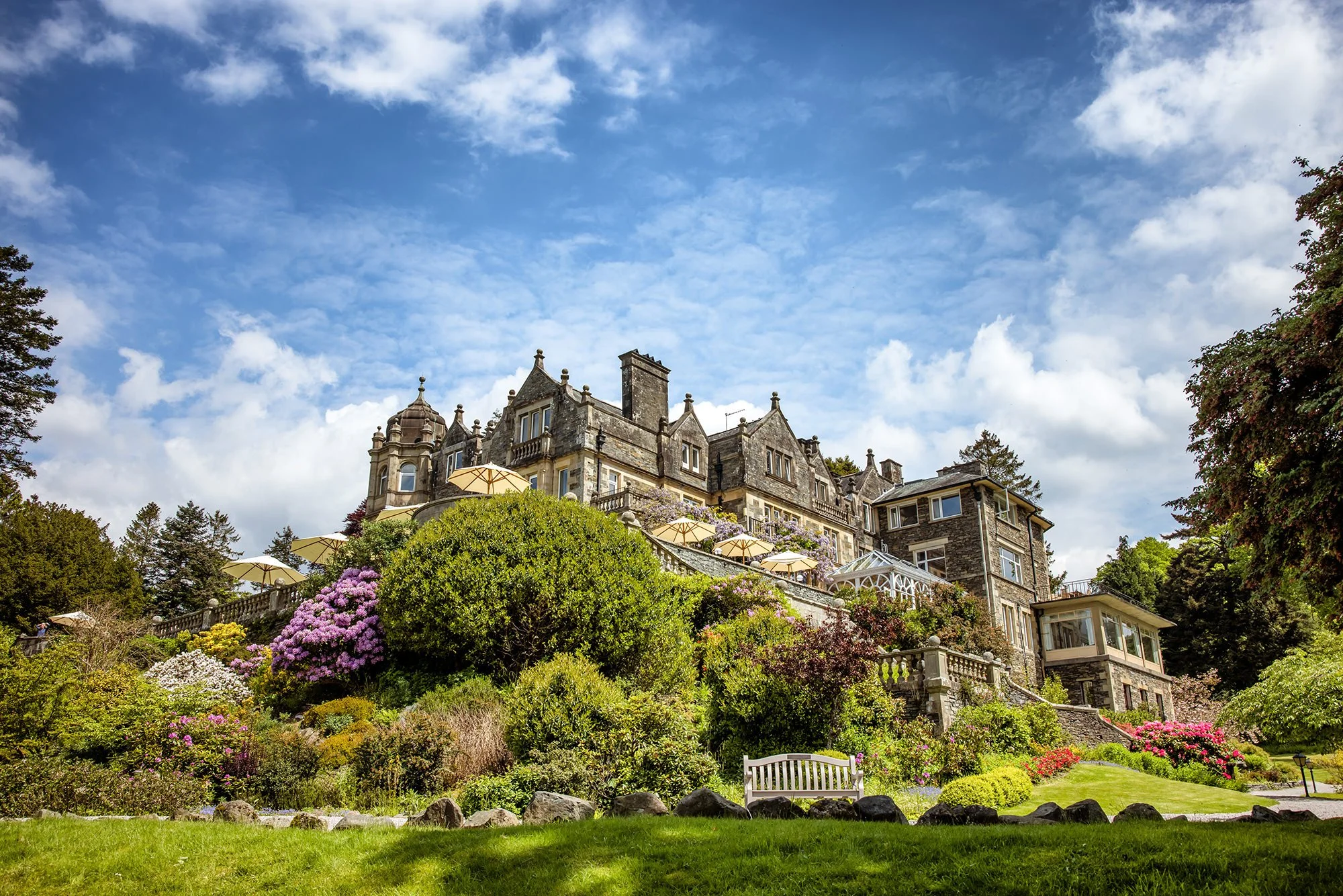 A large, historic stone mansion on a hill, surrounded by lush greenery, colorful flowering bushes, and a well-maintained lawn under a partly cloudy blue sky.
