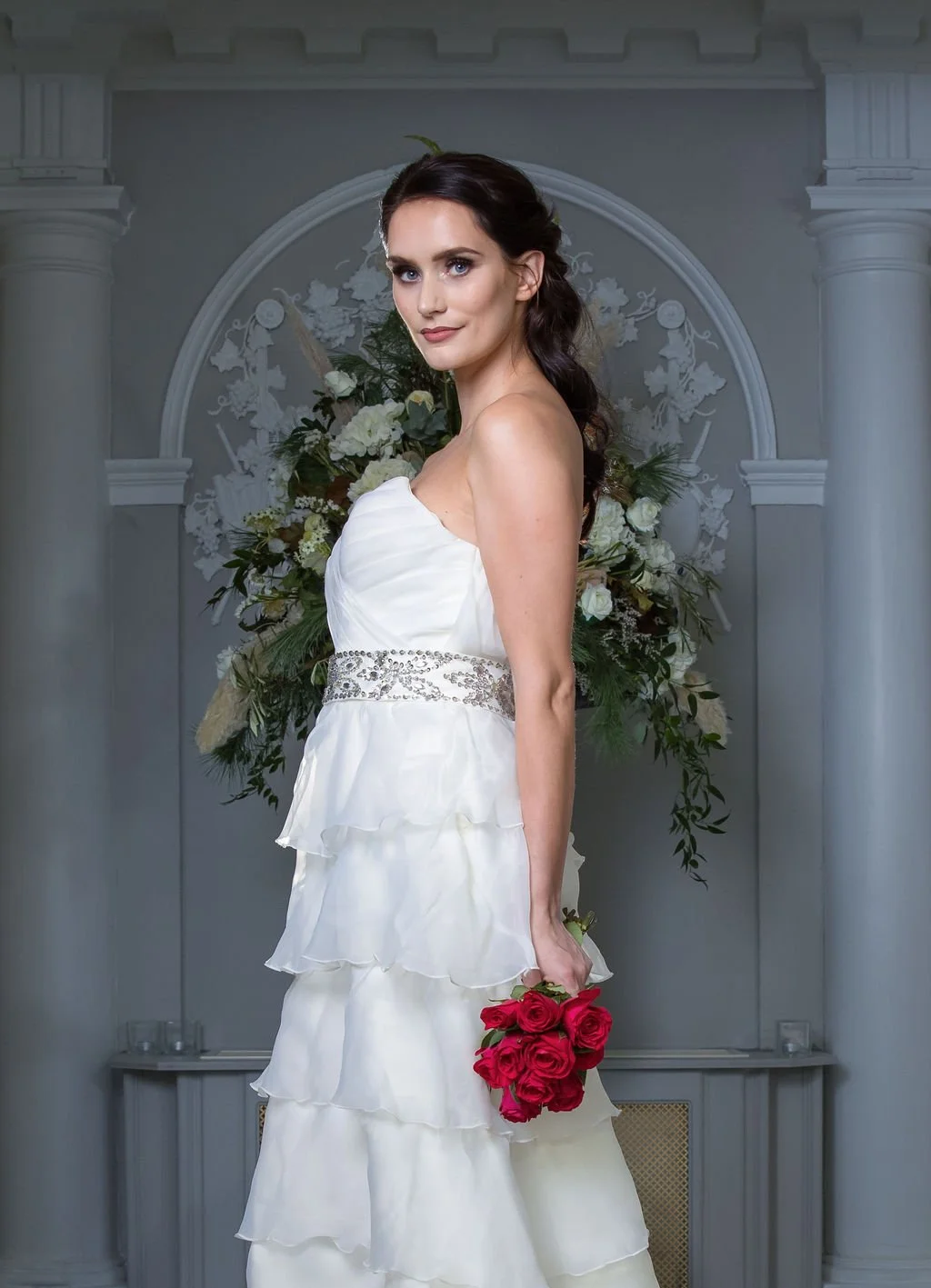 A woman in a white wedding dress holding a bouquet of red roses, standing in front of a floral display with white and green flowers and decor.