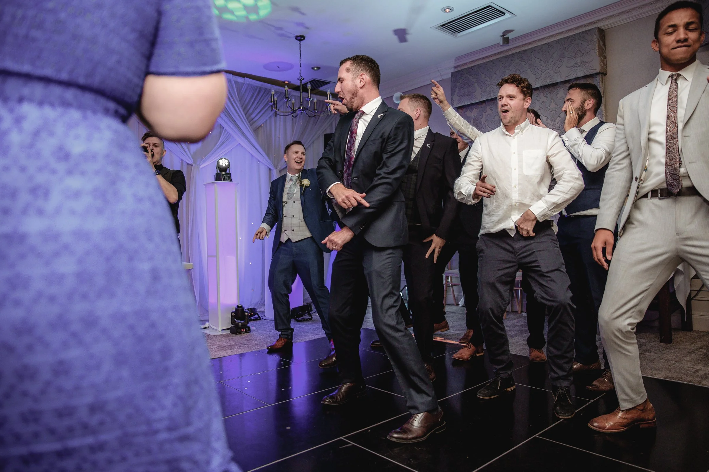 Group of men dancing at a wedding reception in suits and formal attire with purple and white decor in the background.