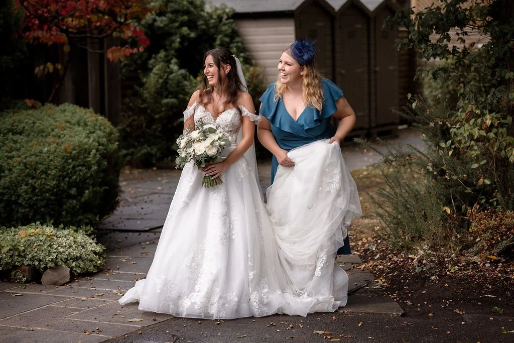 Two women dressed in wedding attire walking outdoors, one holding a bouquet of white flowers and the other lifting her wedding dress, smiling and laughing.