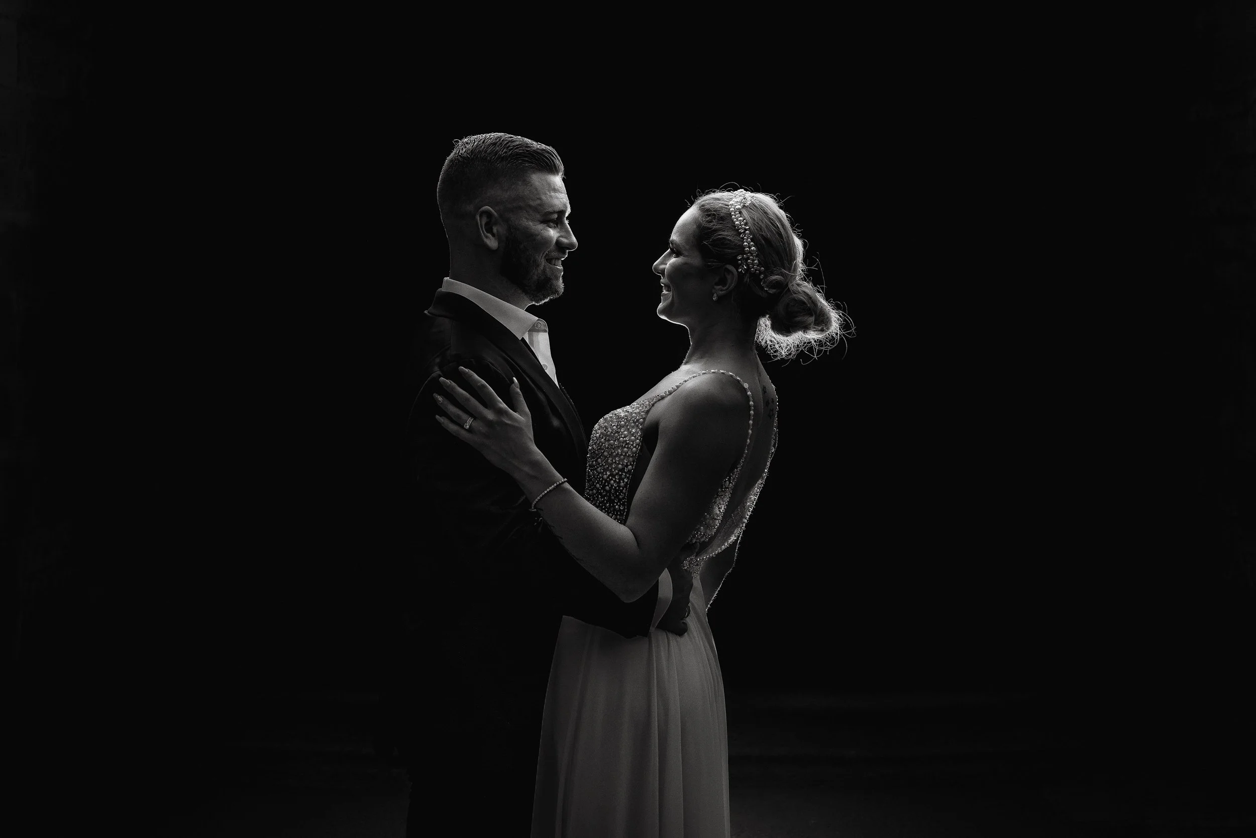 Black and white photo of a bride and groom smiling, standing face to face, with the bride holding the groom's shoulders and the groom's hands around the bride's waist, on a dark background.