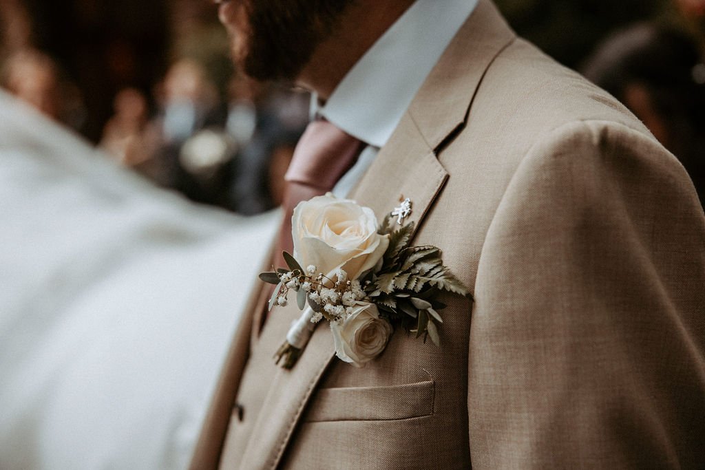 Close-up of a groom's beige suit with a boutonniere of white roses, greenery, and baby's breath