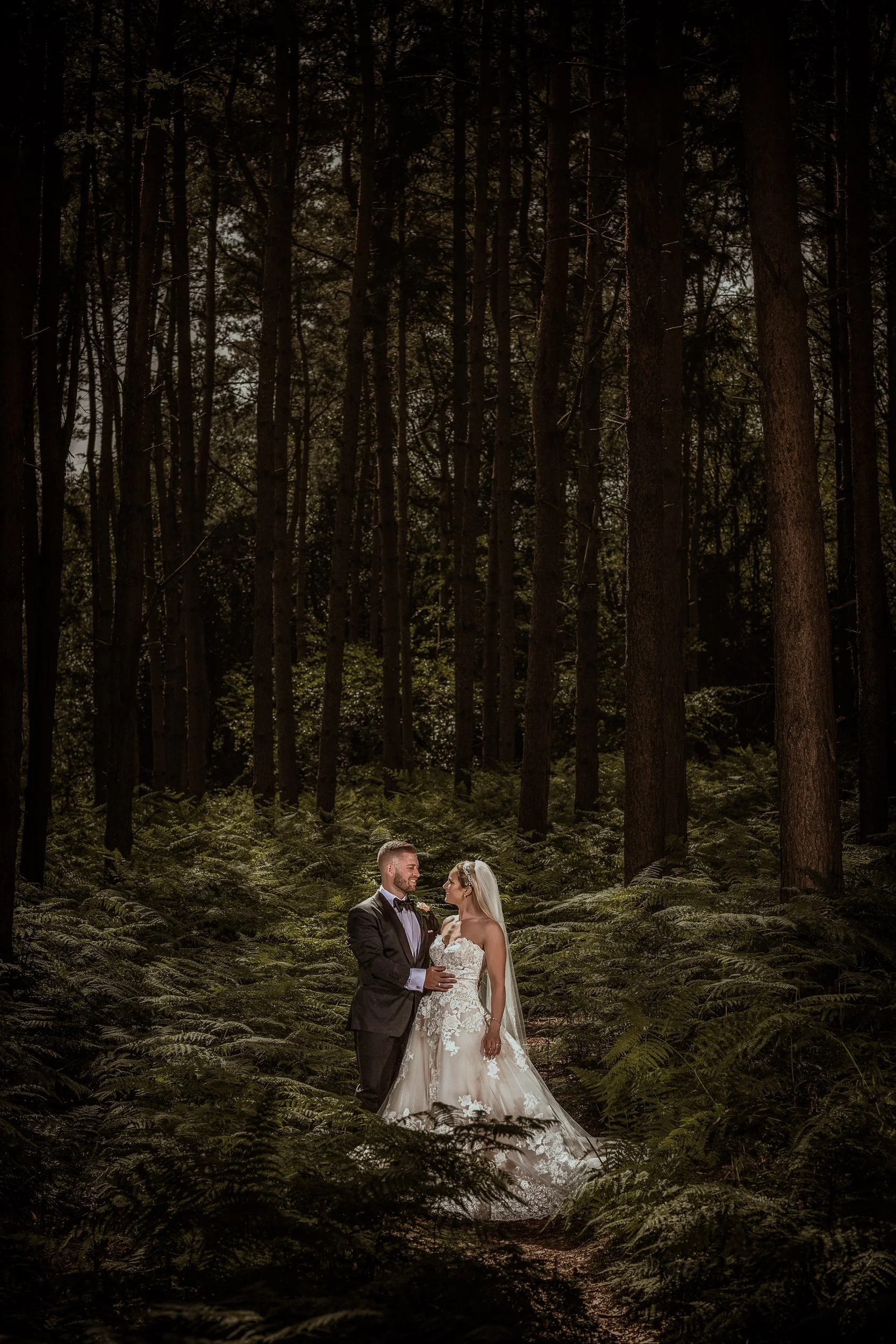 A bride in a white wedding dress and a groom in a black tuxedo stand facing each other in a lush green forest, smiling at each other.