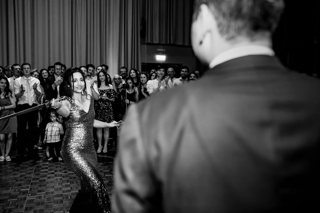 A woman in a sparkling dress is dancing at a formal event, with an audience watching her and clapping.