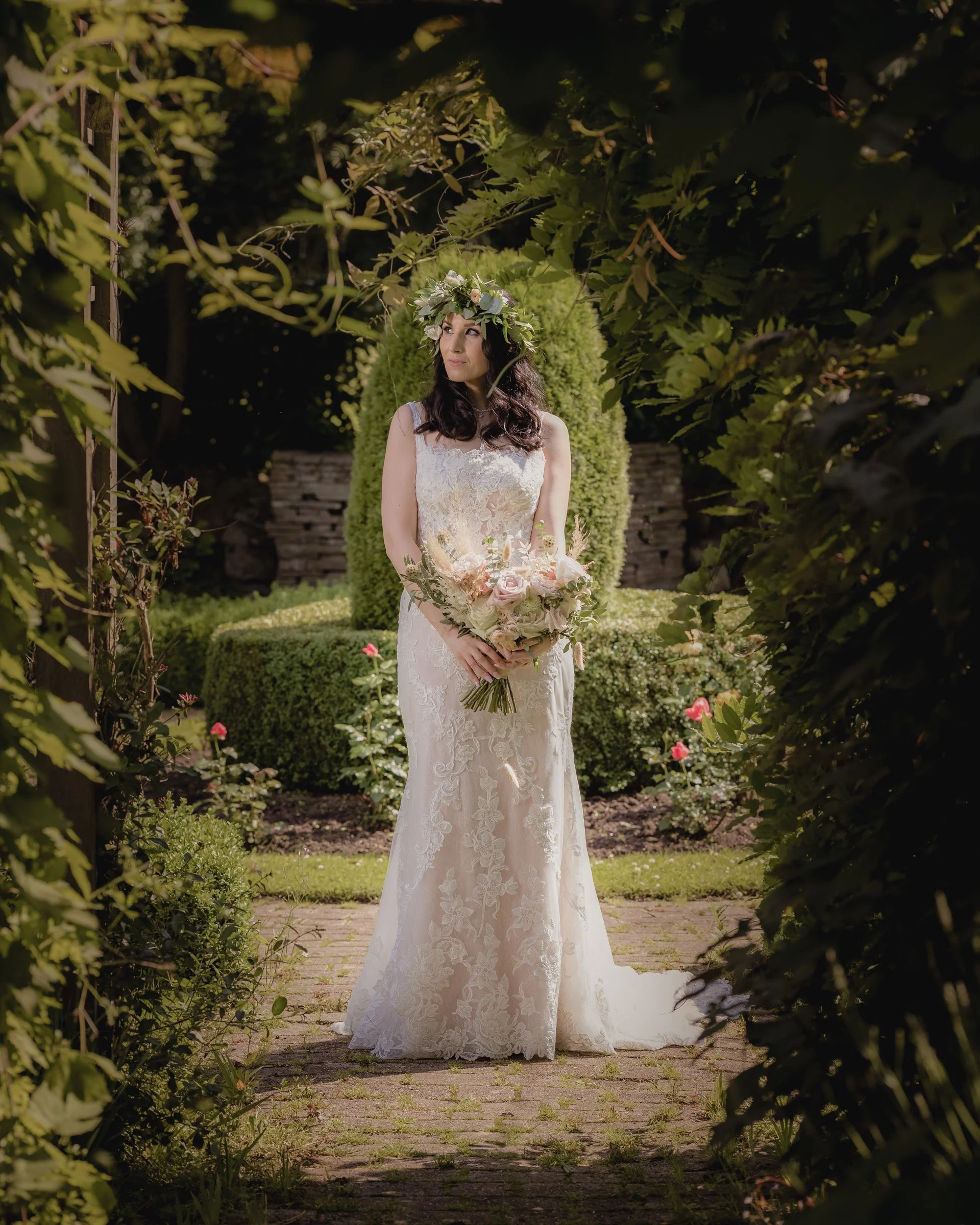 A bride in a white lace wedding gown holding a bouquet of flowers, standing on a brick path in a garden with greenery and pink flowers, framed by leaves.