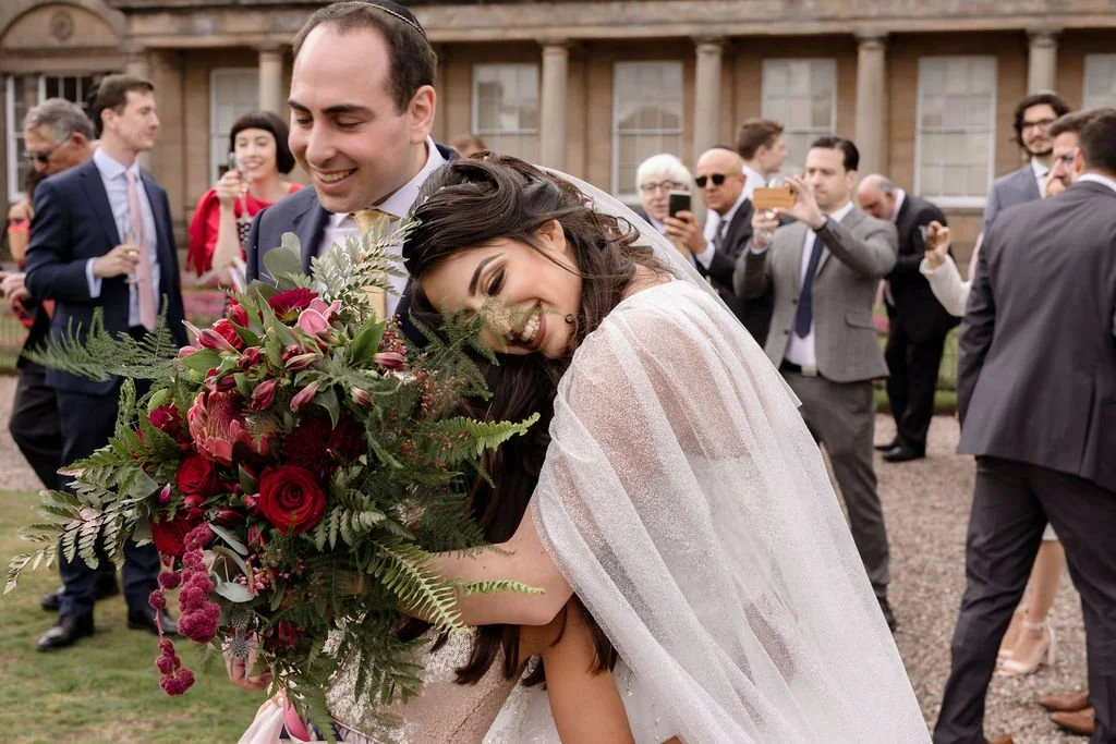 A bride and groom are hugging and smiling at a wedding celebration outdoors, surrounded by guests taking pictures.