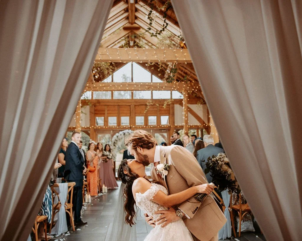 A couple shares a kiss in the center of a rustic wedding venue with wood beams and string lights, surrounded by friends and family.