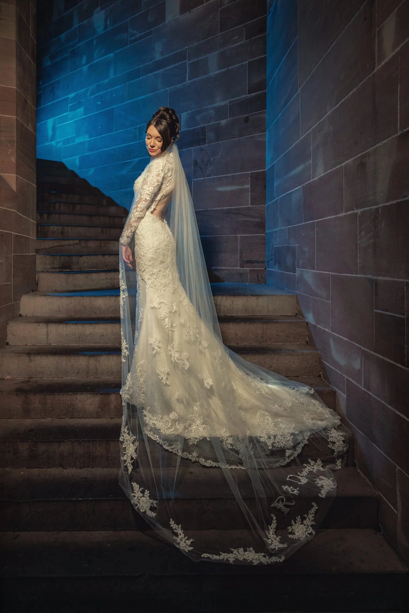 A woman in a long, white bridal gown with lace details and a train, standing on stone stairs against a dark wall with blue lighting.