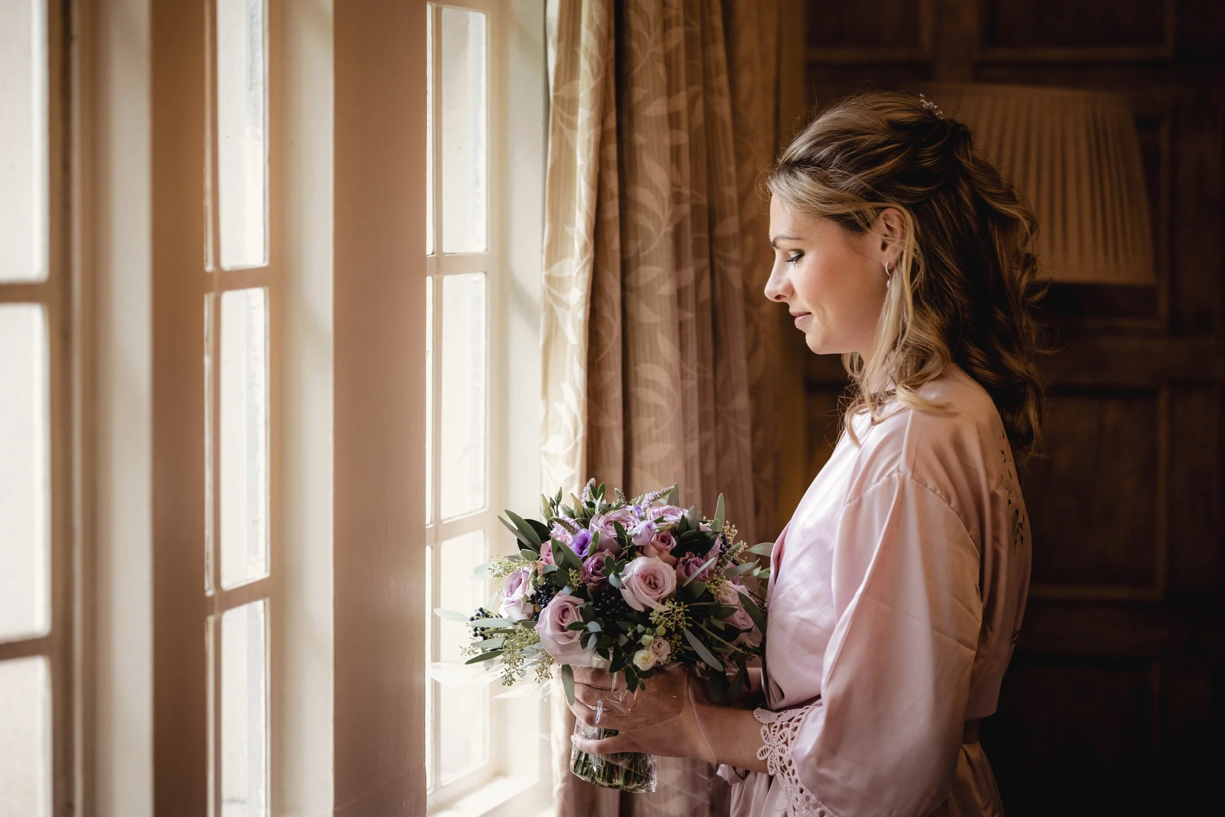 A bride standing by a window, holding a bouquet of pink and purple flowers, dressed in a pink satin robe with lace details, looking down at her bouquet.