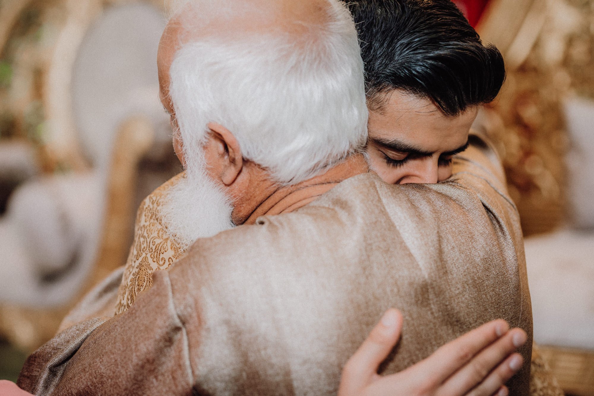 An elderly man with white hair and beard hugging a young man with dark hair, both with closed eyes, in an indoor setting.