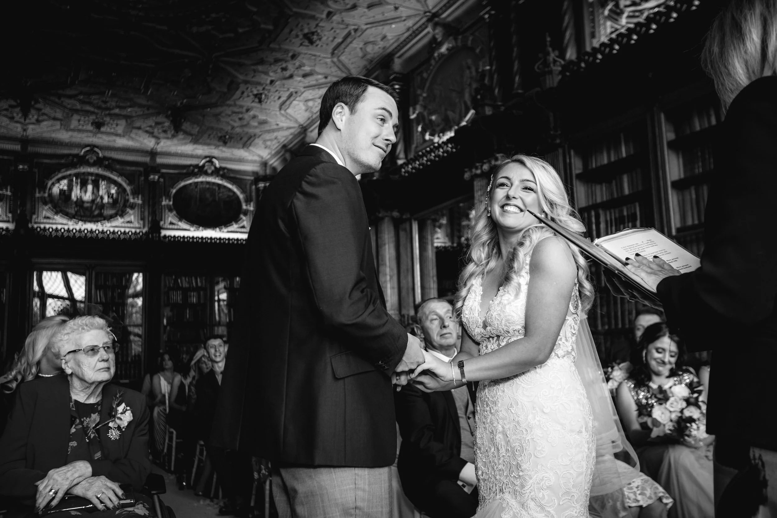 A black-and-white photograph of a wedding ceremony showing a bride and groom holding hands and smiling at each other. The bride is wearing an elegant lace wedding dress and the groom is dressed in a dark suit. Several guests are seated around them, w