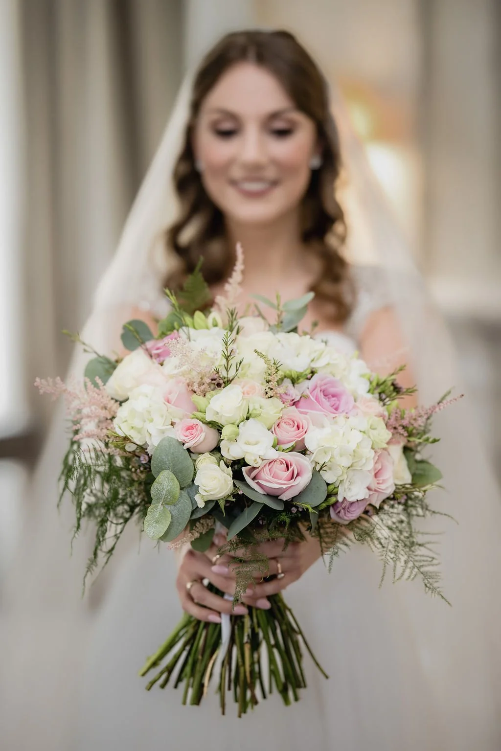 A bride holds a bouquet of pink and white roses, white hydrangeas, and green foliage, smiling softly and wearing a veil and wedding dress.