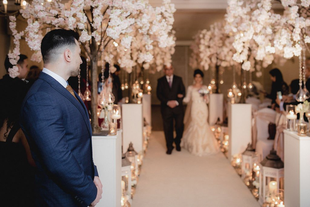 Man in blue suit watching a bride and groom walking down the aisle decorated with white flowers and lanterns.