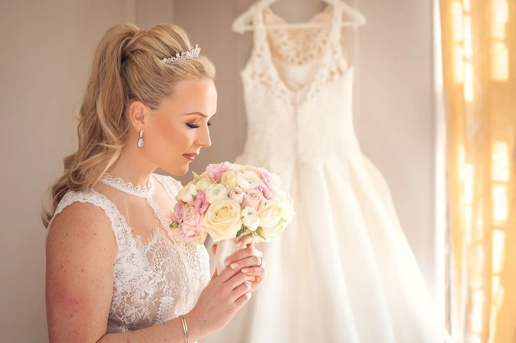 A bride with blonde hair in an updo holding a bouquet of pink and white roses, standing near a wedding dress hanging on the wall.