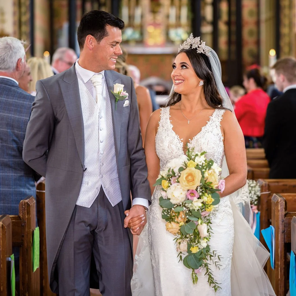 A bride and groom holding hands, smiling at each other during their wedding ceremony inside a church with guests in the background.