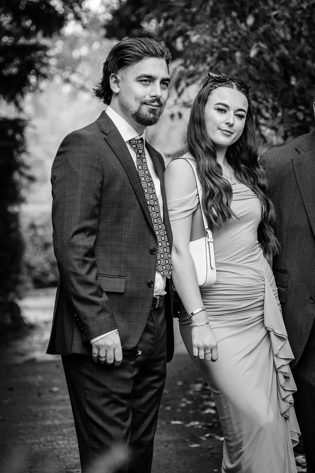 A black and white photo of a man and a woman standing outdoors, dressed formally, with trees in the background.