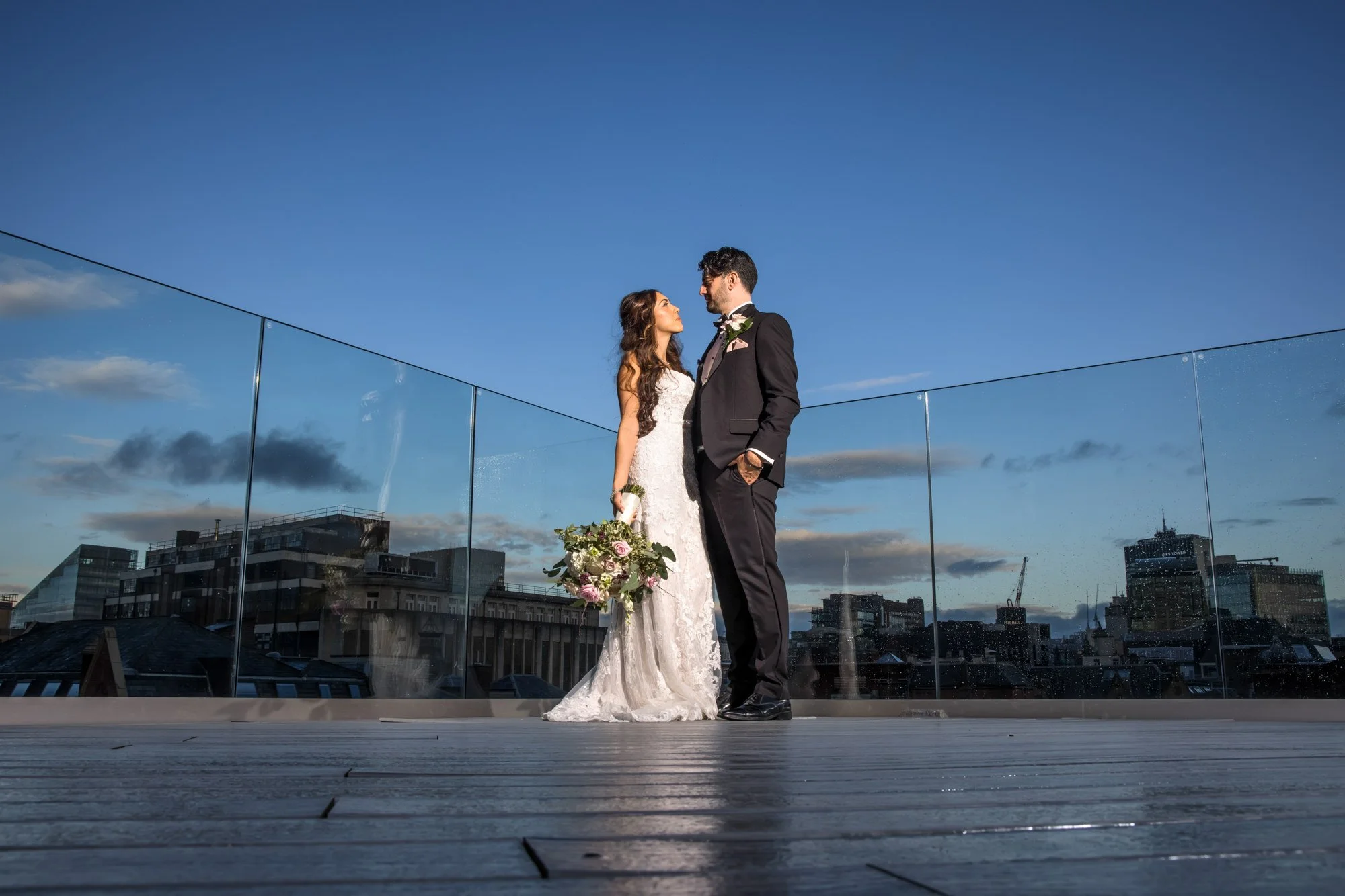 A bride and groom stand closely on a rooftop terrace, gazing into each other's eyes with the cityscape and a blue sky with clouds in the background.