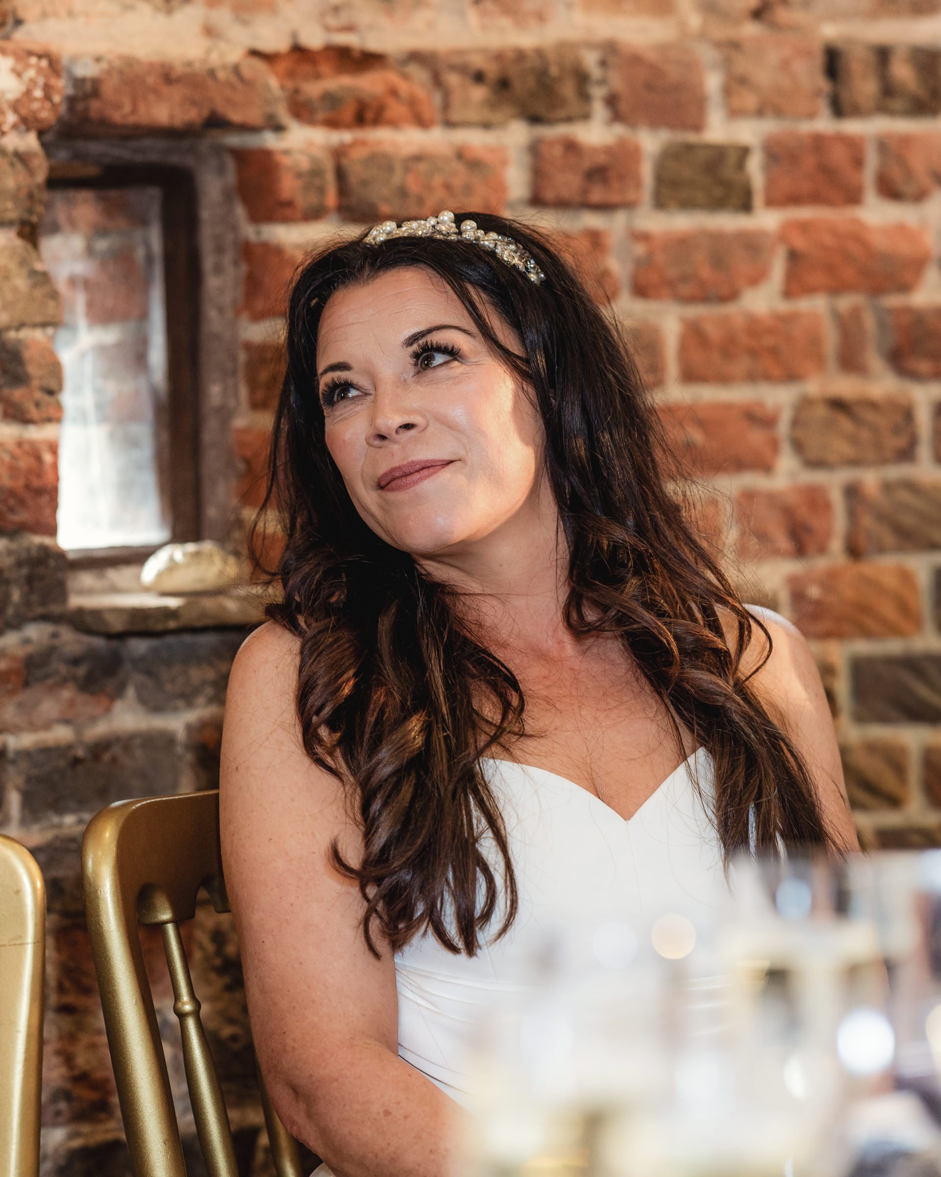 A woman with dark wavy hair wearing a white dress and a pearl headband, sitting inside near a brick wall with a small window.