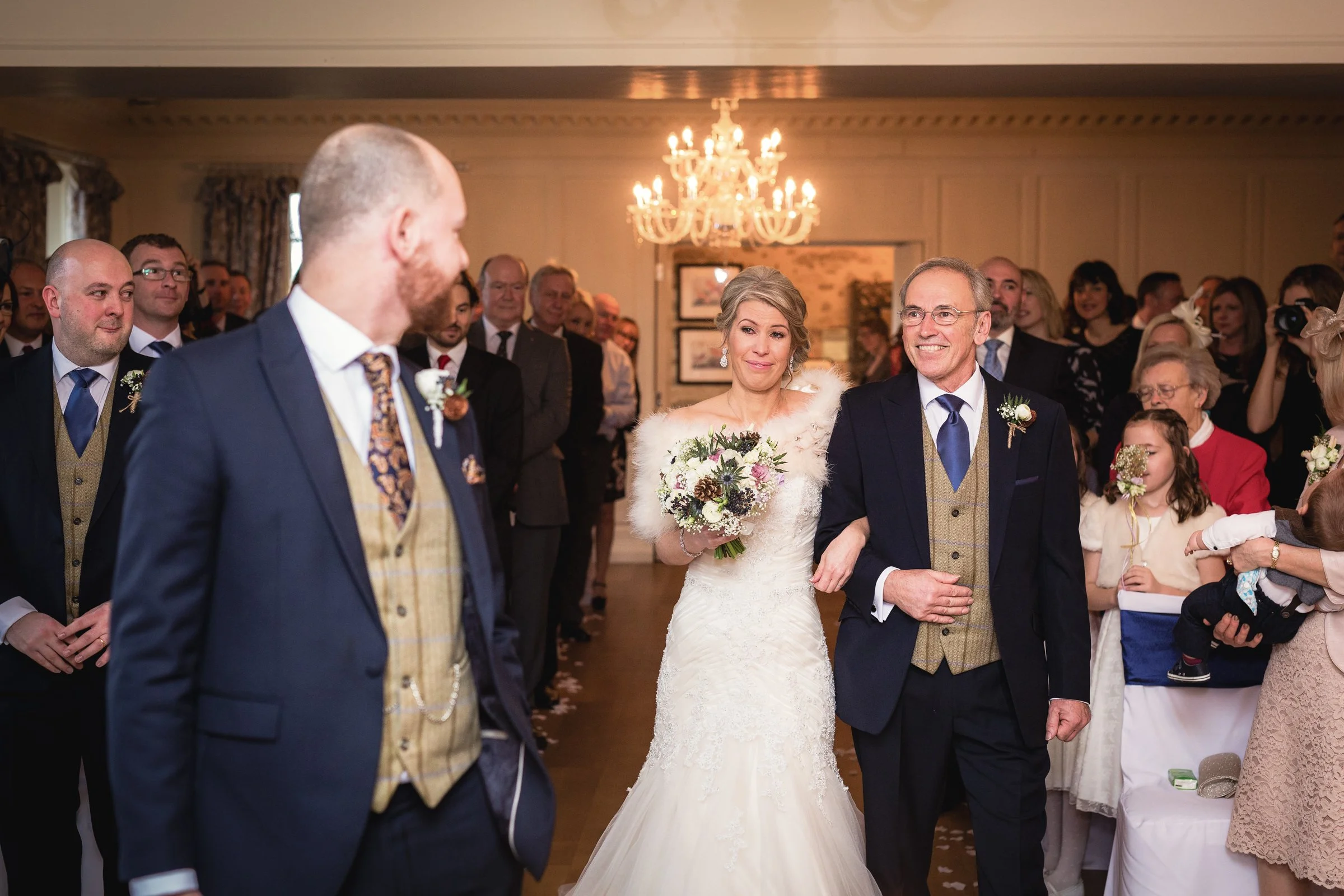 A bride walking down the aisle arm-in-arm with an older man, likely her father, at a wedding ceremony. The bride wears a white wedding gown and holds a bouquet, while the man wears a dark suit with a beige vest. Guests are seated and standing, watchi