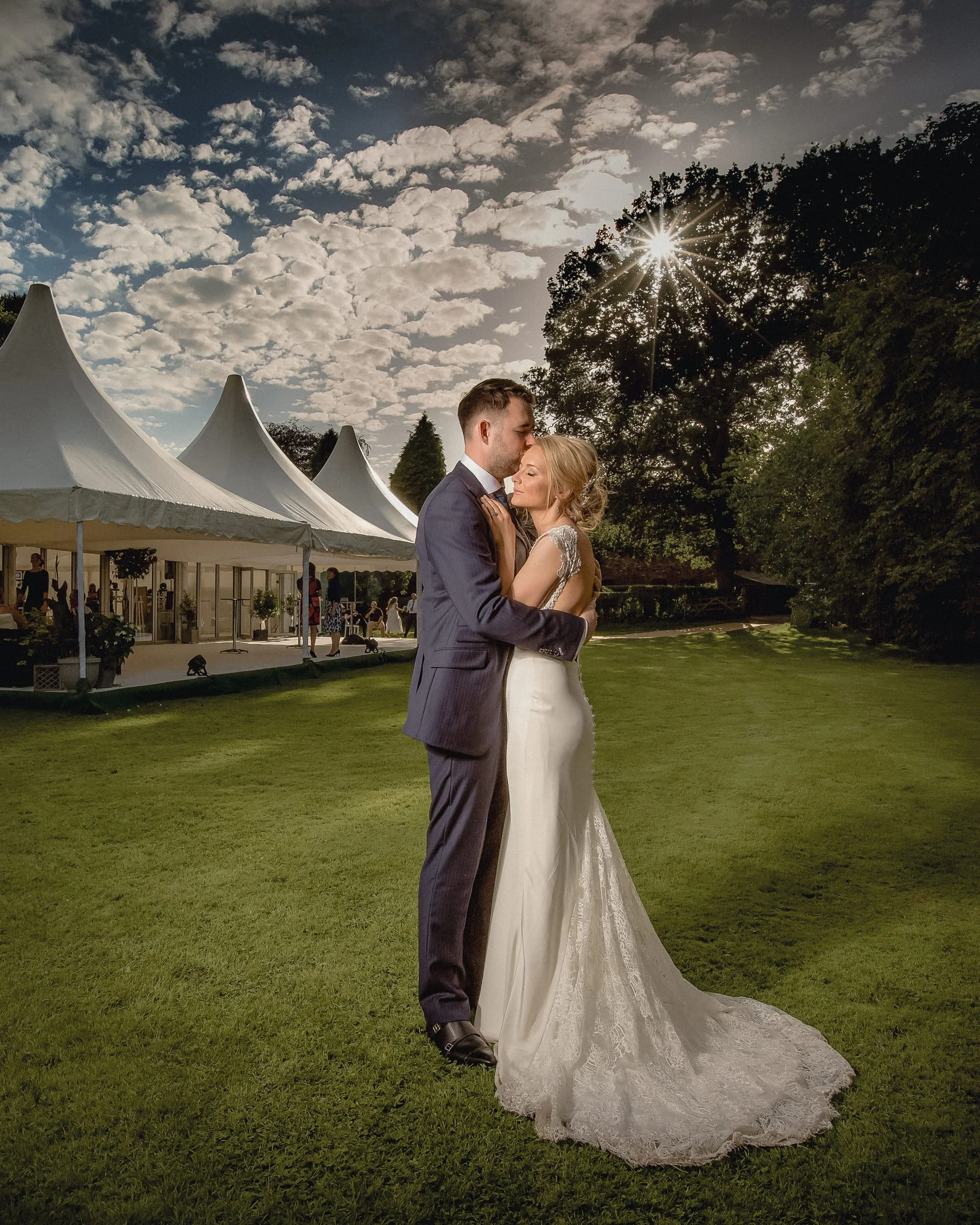 A bride and groom share a kiss outdoors at sunset, with a festival tent and trees in the background.