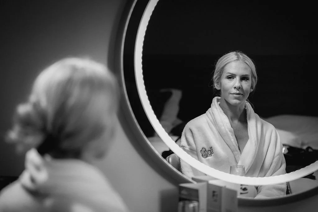 A woman looking at herself in a circular mirror, wearing a bathrobe with a logo on it, in a room with a bed in the background.