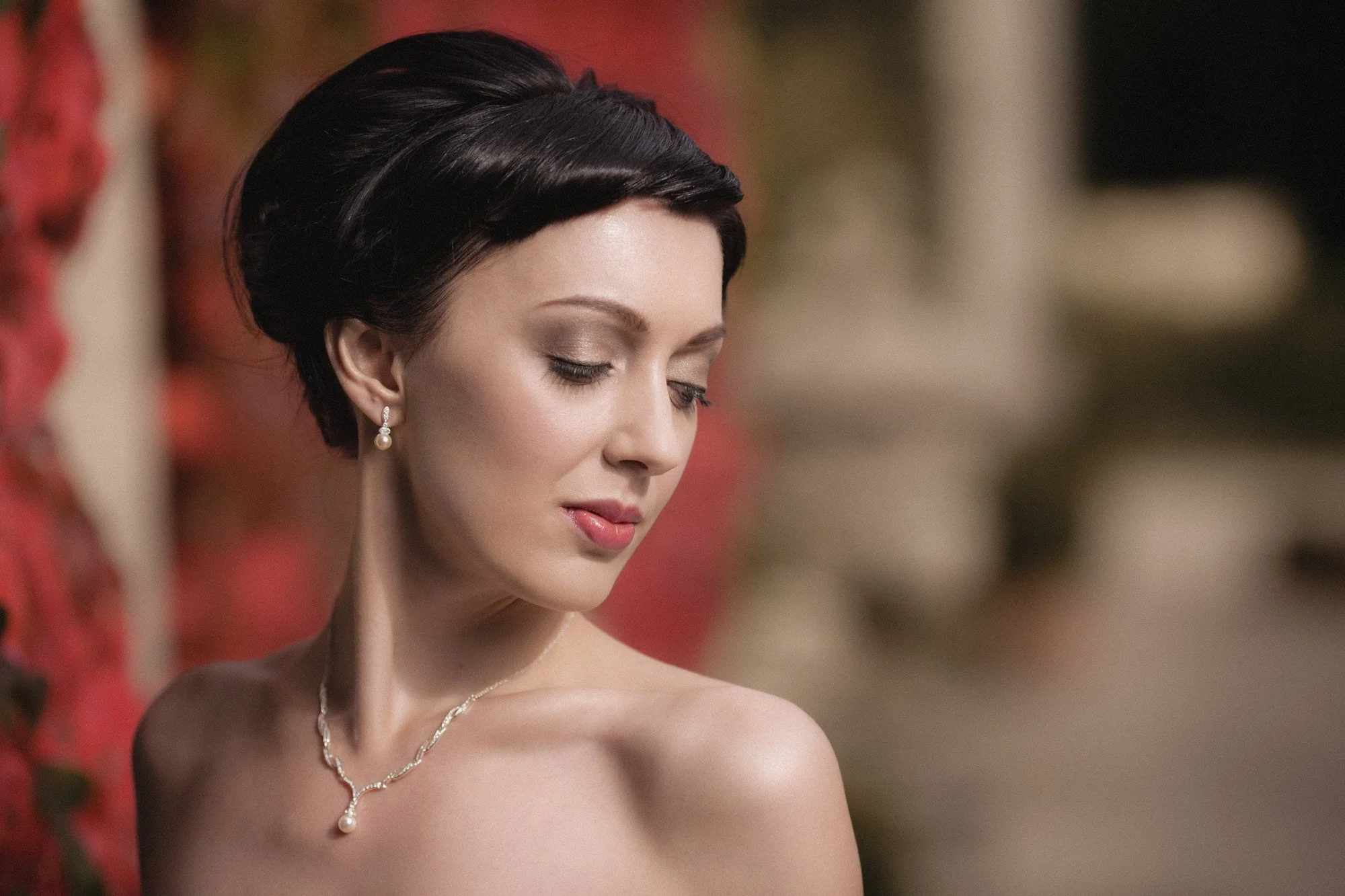 A woman with short dark hair, wearing pearl earrings and a pearl necklace, looking downward with a soft expression, against a blurred background.