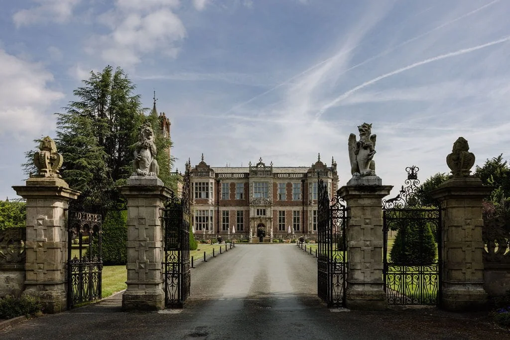 A large, historic mansion with intricate architecture is viewed behind an ornate black wrought iron gate topped with stone lion sculptures. Tall trees surround the mansion, and a clear, partly cloudy sky is overhead.